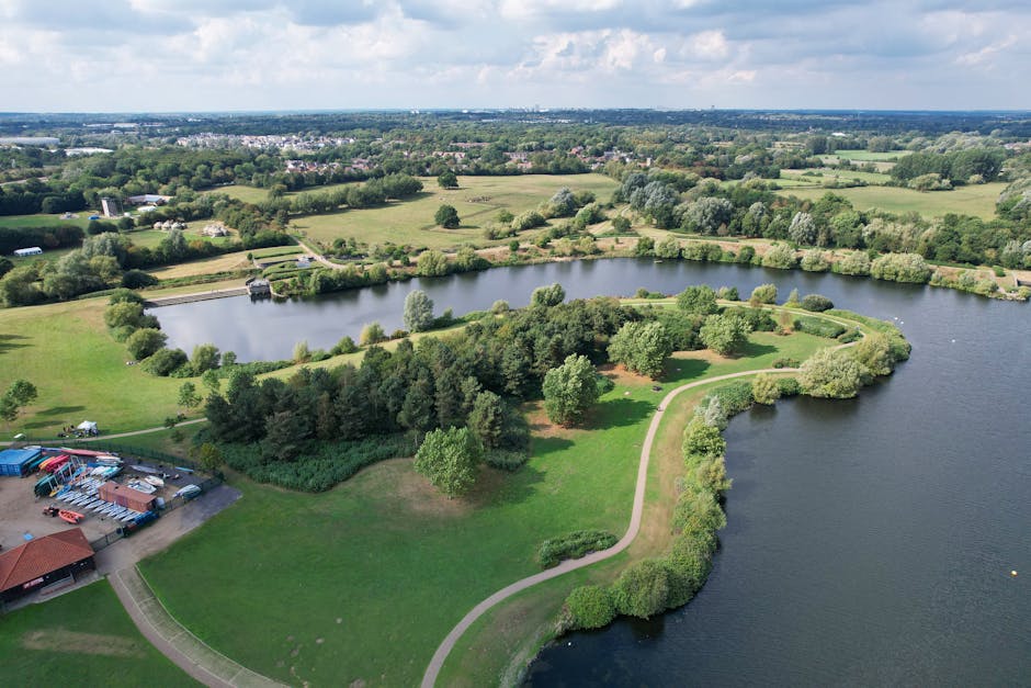 An aerial view of Rickmansworth Aquadrome featuring a large, irregularly shaped lake with calm, dark blue waters bordered by a narrow, winding pathway. The pathway, visible from the bottom right, curv