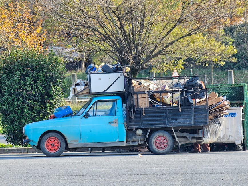 A roadside scene featuring an overflowing pile of mixed household rubbish and waste materials situated on a gravel surface in front of a low stone wall. Visible objects include black plastic bin bags 