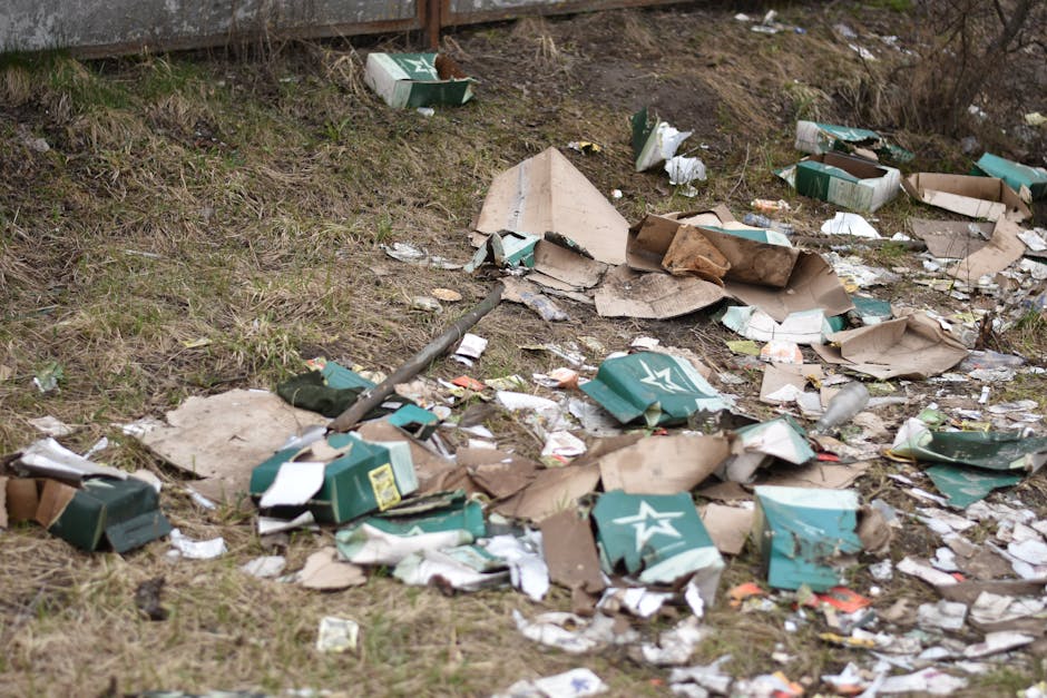 A close-up image showing a large collection of crumpled aluminium cans and crushed beverage bottles, mainly silver, with some red, blue, yellow, and green labels, scattered and overlapping each other 